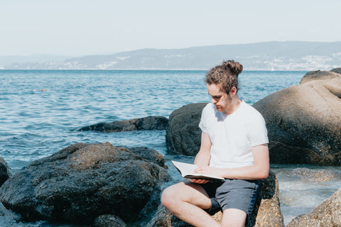 Mann sitzt auf Steinen am Meer und liest